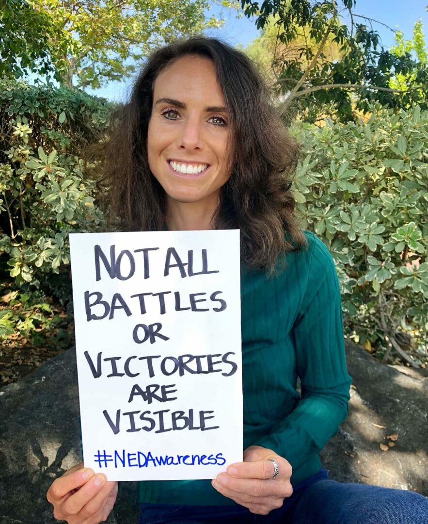 Brittany is holding a sign that reads "Not all battles or victories are visible. #NEDAwareness"