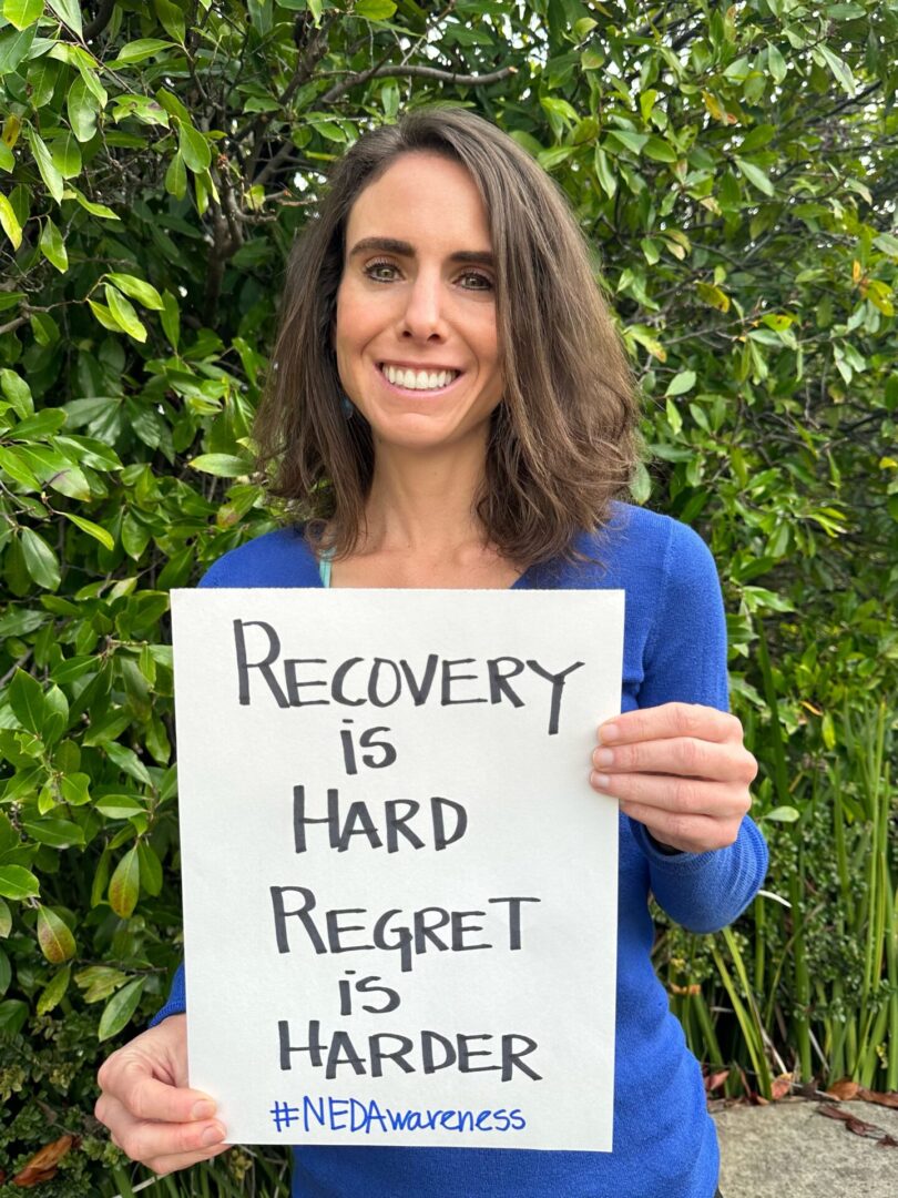 Brittany is holding a sign for NEDA awareness week that reads "Recovery is hard. Regret is harder."